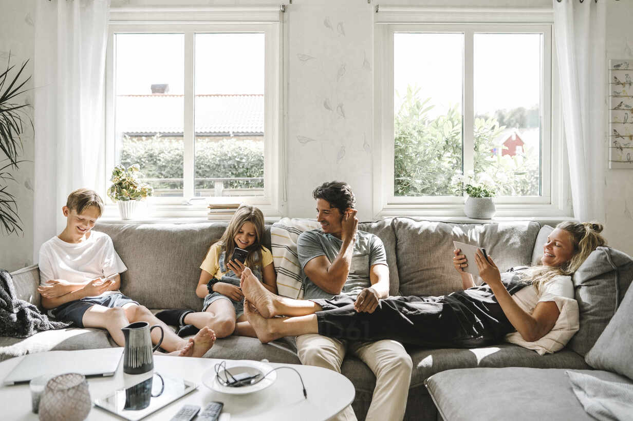 A family of four relaxes on a couch in a bright living room, each seemingly engaged with their own devices. This could represent a dynamic explored in family therapy counseling, such as communication or individual needs within the family unit.