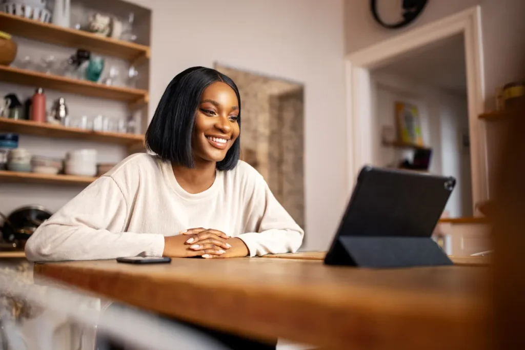 A smiling Black woman with short dark hair looks at a tablet screen, participating in an online counseling session.