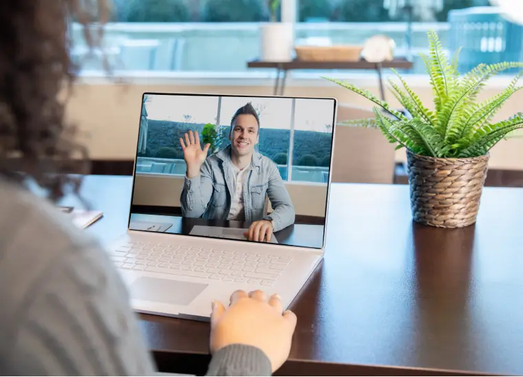 A person views a laptop screen showing another individual waving during an online counseling session. The laptop sits on a wooden table with a potted plant nearby.