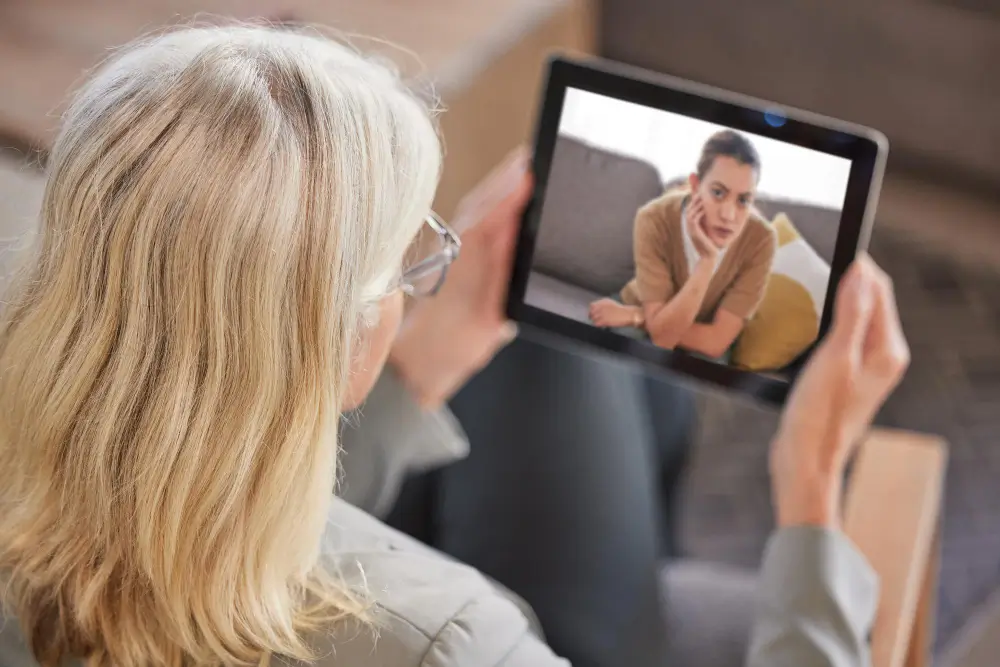 A woman engages in an online counseling session via tablet, looking at the image of another woman on the screen.